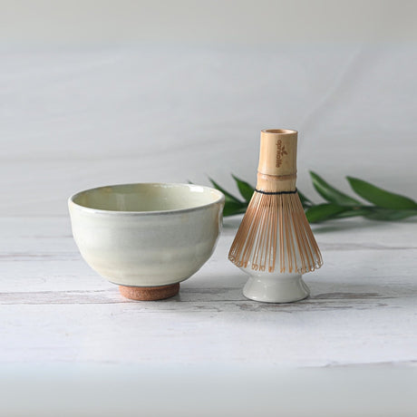 A serene setting shows a Ceramic Matcha Tea Starter Set on a white surface, lush greenery behind. The White Kohiki Ceramic Matcha Bowl, crafted with Minoyaki pottery techniques, adds authenticity alongside the bamboo matcha whisk.