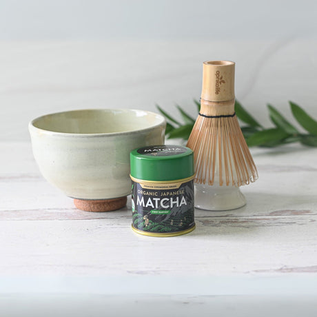 White Kohiki Ceramic Matcha Bowl Set with matcha tin and bamboo whisk on a light wooden surface, surrounded by leaves.