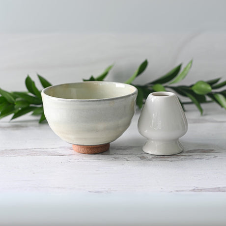 The White Kohiki Ceramic Matcha Bowl rests on a light wooden surface, accompanied by a small vase crafted with Minoyaki techniques, while green foliage sets a serene background.