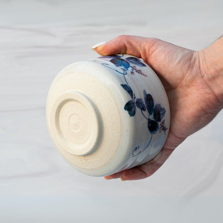A hand holds an upside-down ceramic bowl with a blue floral design against a light background. This elegant piece, part of the White Clay Kudzu Flowers Matcha Bowl, Bamboo Matcha Whisk, Whisk Holder and Organic Japanese Matcha Set, is crafted in the renowned Minoyaki pottery tradition.