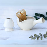 The White Ceramic Matcha Bowl with Spout, accompanied by a bamboo whisk and a small white vase on a wooden surface, makes an ideal housewarming gift.