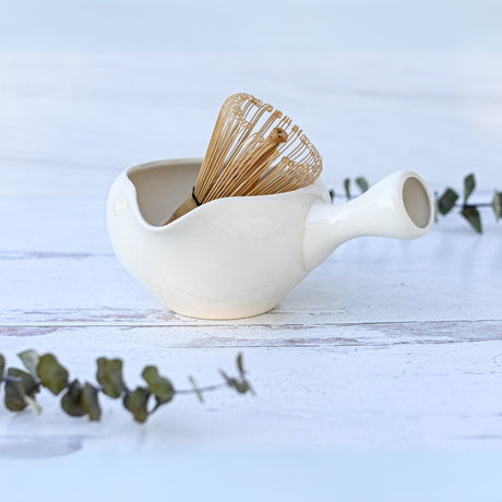 The White Ceramic Matcha Bowl with Spout, along with a bamboo whisk, is perfect for a Japanese tea ceremony and sits gracefully amid eucalyptus leaves on a light wooden surface.