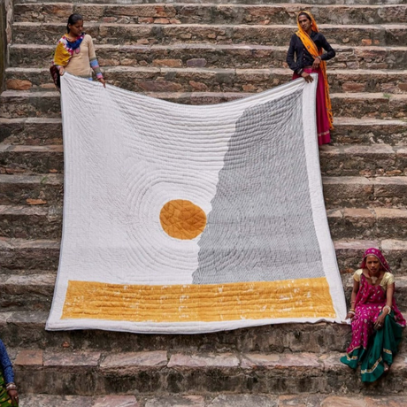 People holding a large knitted blanket with a sun design on stone steps.