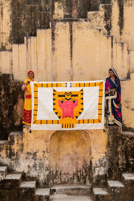 Two women holding a colorful quilt with a tiger design against an old stone wall.