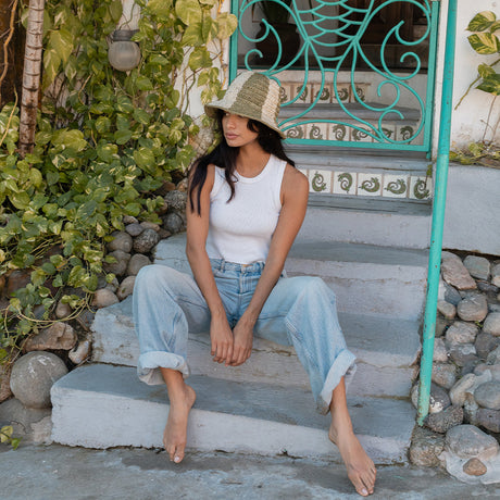 Woman sitting on steps wearing a white tank top and light blue jeans, with a straw hat.