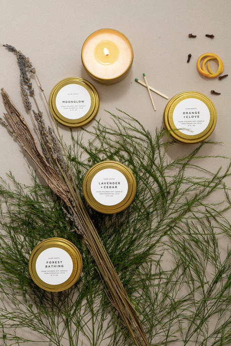 Five candles in gold tins with labels on a neutral background with greenery and wooden sticks.