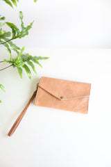 A brown leather wallet with a brass snap and a leather wristlet, displayed on a white background with a green plant leaf partially visible to the left.