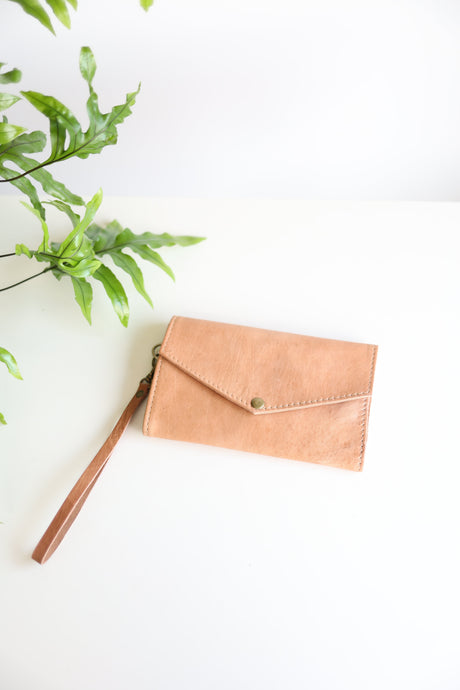 A brown leather wallet with a brass snap and a leather wristlet, displayed on a white background with a green plant leaf partially visible to the left.