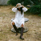 Woman in a white dress and brown boots sitting on a wooden stool outdoors.