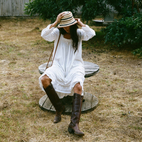Woman in a white dress and brown boots sitting on a wooden stool outdoors.