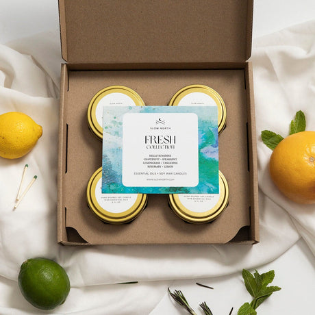 Box of candles with a card labeled 'Fresh Collection' on a white fabric background with fruits and herbs.