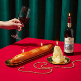 Hand holding a glass of red wine with a bottle, bread, and candles on a red tablecloth against a green curtain.