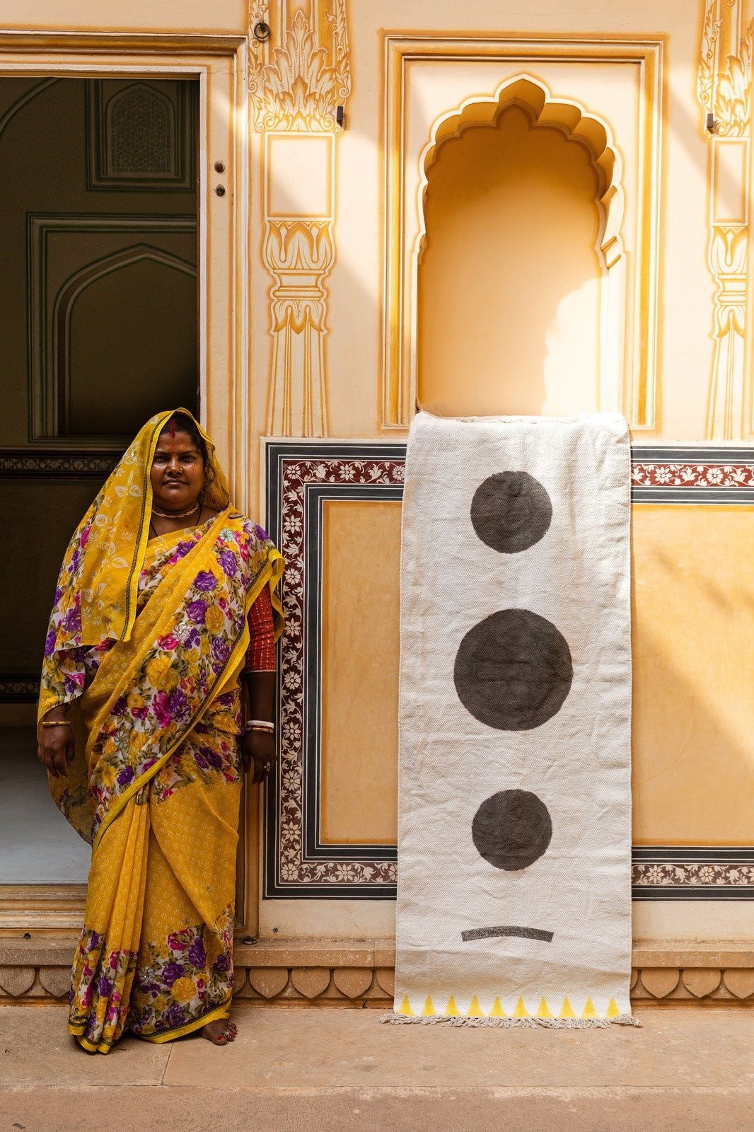 Woman in a yellow saree standing next to a decorative wall panel with circular patterns.