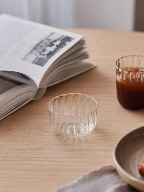 Clear glass cup on a wooden table with an open book and a small bowl in the background.