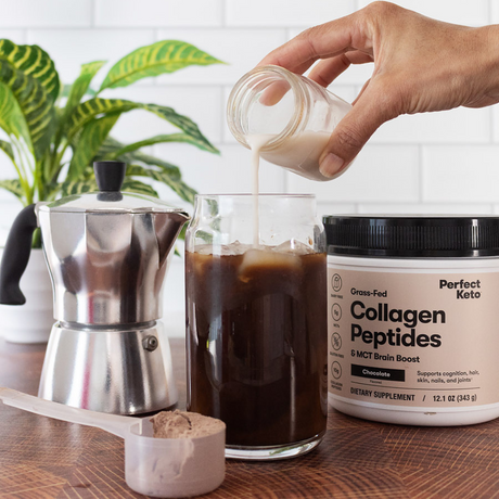 Person pouring a liquid into a glass of coffee with a container of Perfect Keto Collagen Peptides on a wooden surface.