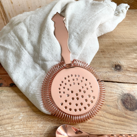 Rose gold cocktail strainer on a wooden surface with a white cloth