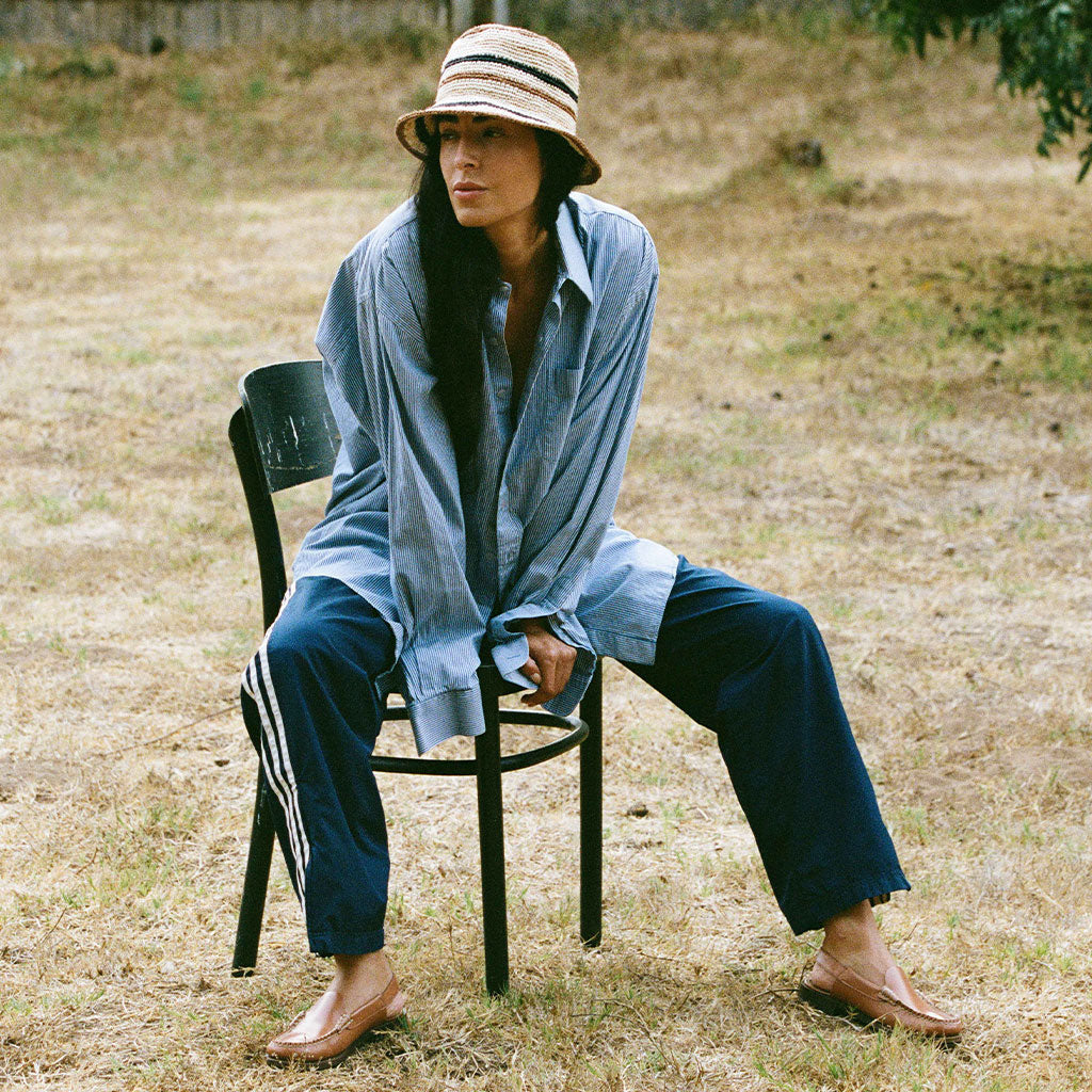 Person sitting on a chair outdoors wearing a blue shirt and pants with a straw hat.
