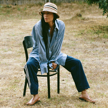 Person sitting on a chair outdoors wearing a blue shirt and pants with a straw hat.