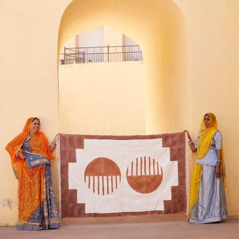 Two women holding a large decorative rug with geometric patterns against a beige wall.
