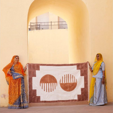 Two women holding a large decorative rug with geometric patterns against a beige wall.