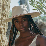 Woman wearing a beige hat with braided hair, standing outdoors with trees in the background