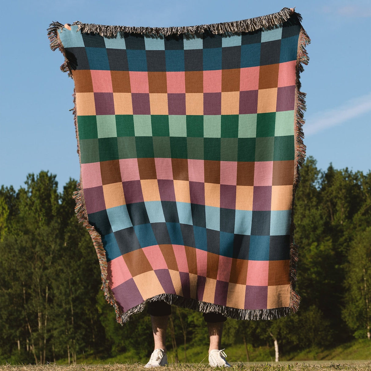 Person holding a colorful checkered blanket outdoors with trees and blue sky in the background
