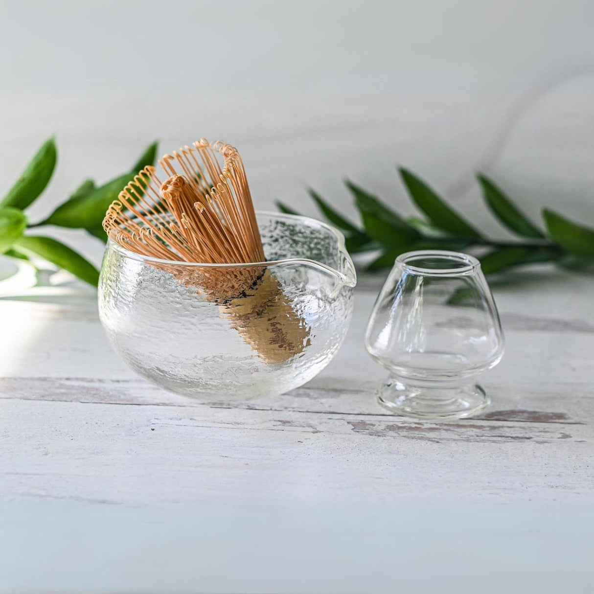 The Textured Spouted Glass Matcha Bowl Pattern 1, with a bamboo matcha whisk and whisk holder set, rests elegantly beside a small cup on a white surface, backed by vibrant green leaves—an ideal housewarming gift for those who appreciate serene aesthetics.