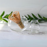 The Textured Spouted Glass Matcha Bowl Pattern 1, with a bamboo matcha whisk and whisk holder set, rests elegantly beside a small cup on a white surface, backed by vibrant green leaves—an ideal housewarming gift for those who appreciate serene aesthetics.