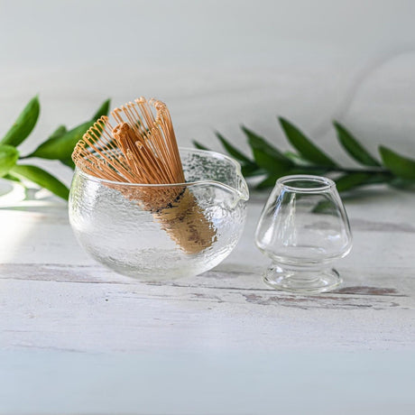 The Textured Spouted Glass Matcha Bowl Pattern 1, with a bamboo matcha whisk and whisk holder set, rests elegantly beside a small cup on a white surface, backed by vibrant green leaves—an ideal housewarming gift for those who appreciate serene aesthetics.