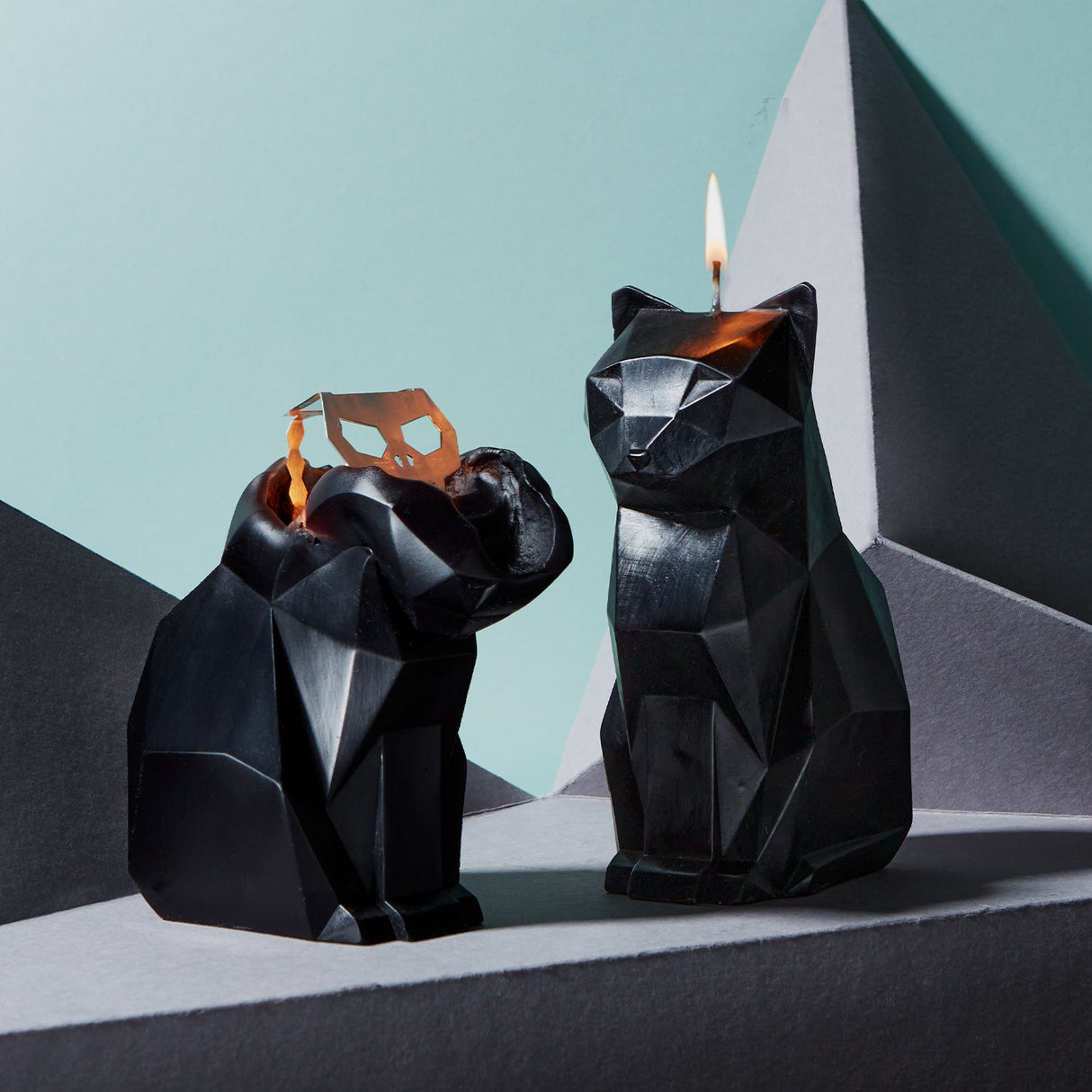 Two black geometric cat-shaped candle holders with lit candles against a light blue and gray background.