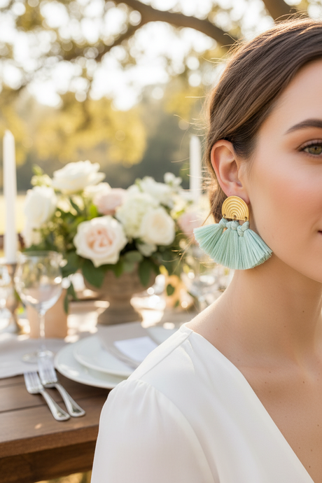 Woman wearing light blue tassel earrings with a blurred outdoor setting featuring flowers and a table.
