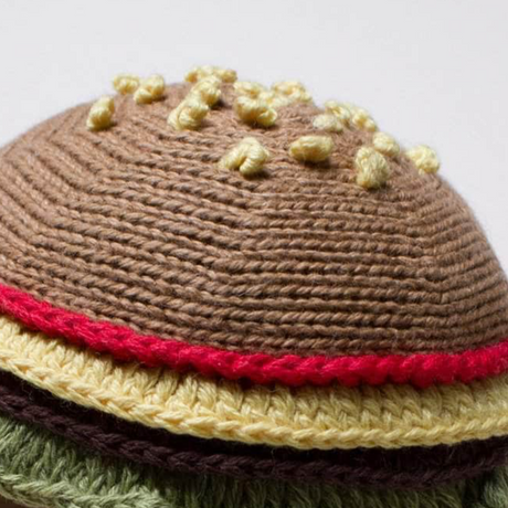 Knitted toy resembling a hamburger with brown bun, yellow cheese, red tomato, and green lettuce on a white background.