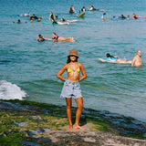 Woman in a yellow bikini top and denim shorts standing on a beach with people swimming and surfing in the ocean.