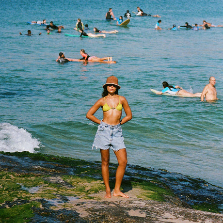 Woman in a yellow bikini top and denim shorts standing on a beach with people swimming and surfing in the ocean.